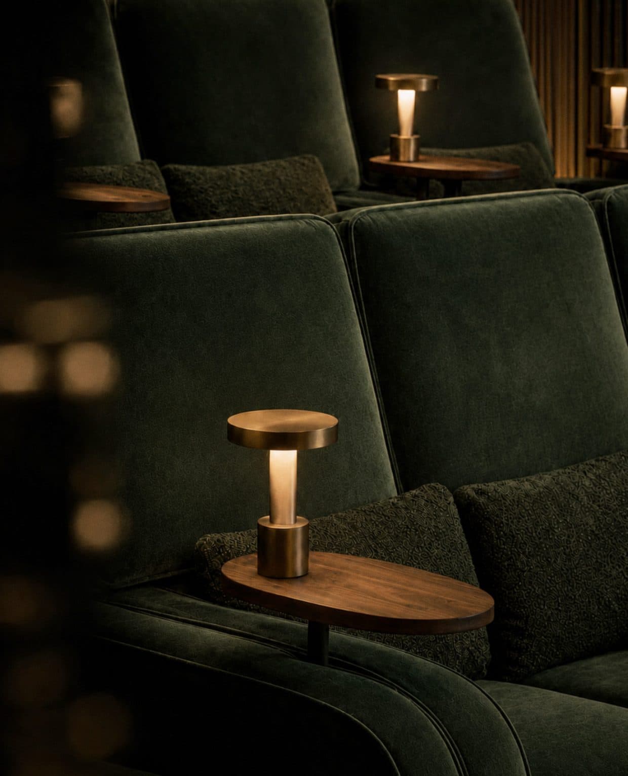 Rows of deep green velvet cinema seats with cushions, each with a small walnut side table and a brass table lamp, in a dimly lit room.