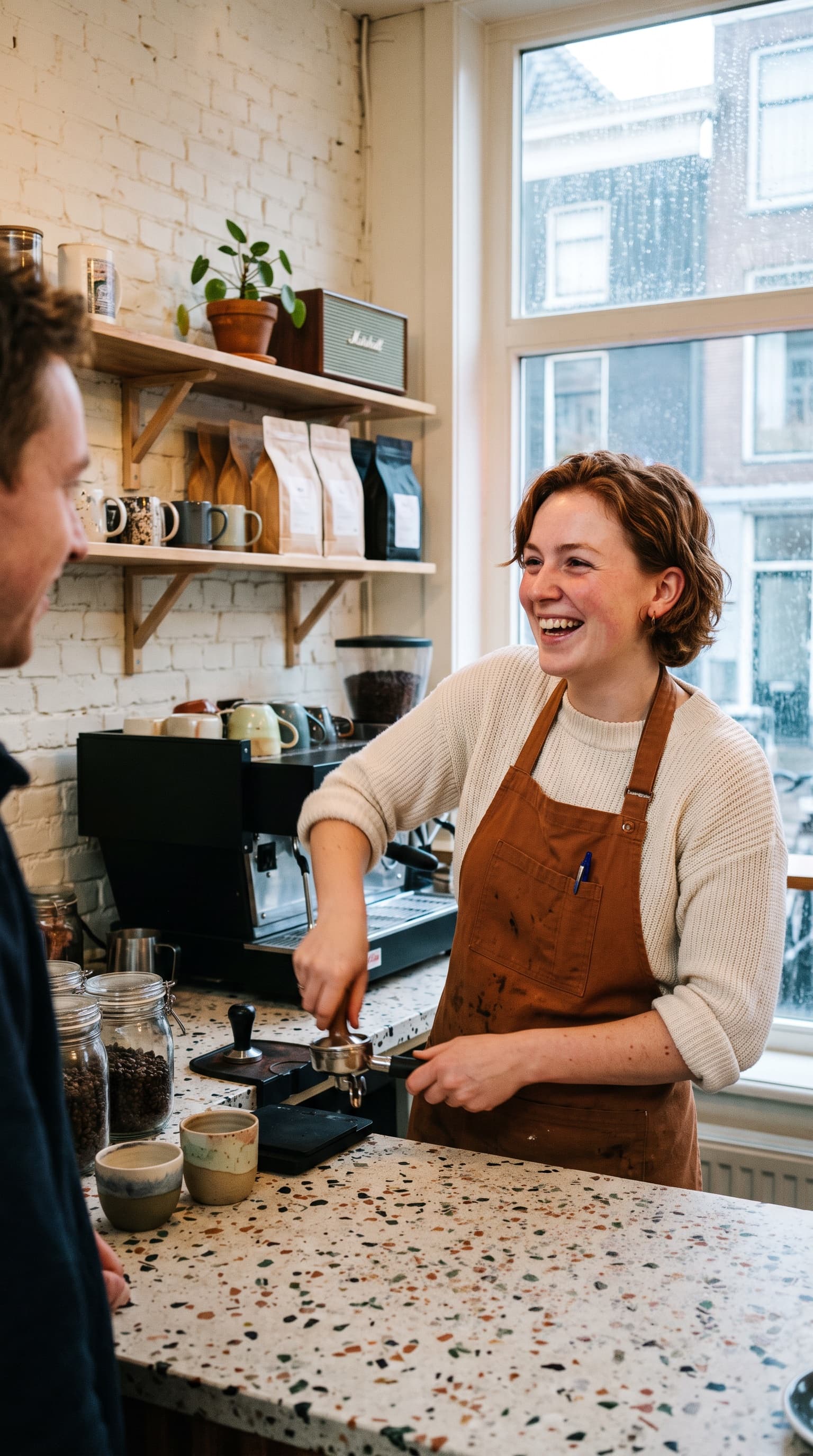 A laughing barista in a brown apron tamping espresso behind a terrazzo counter, shelves of cups and coffee bags against a white brick wall, a tall window onto the street.
